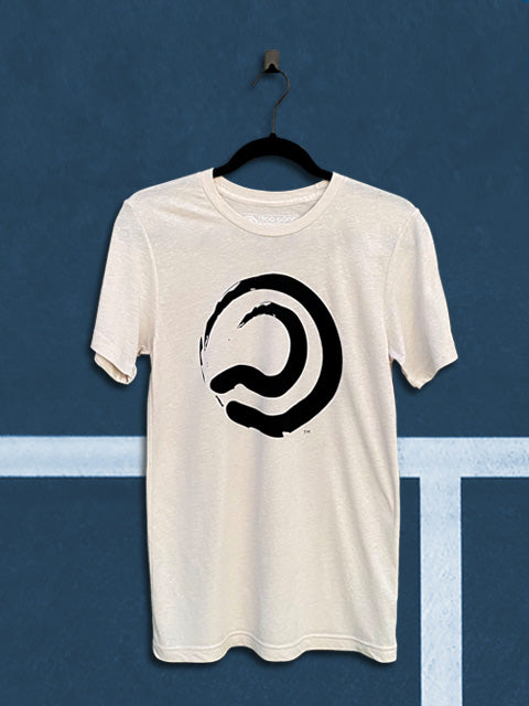 Cement colored t-shirt with a black ball mark graphic, hanging from a black hanger in front of a teal tennis court background.