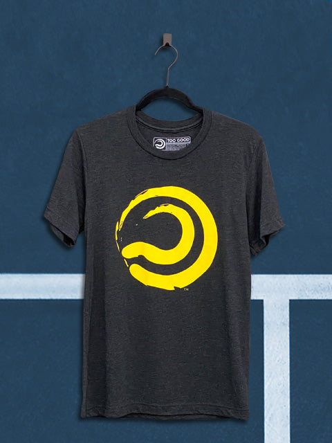 Charcoal colored t-shirt with a yellow ball mark graphic, hanging from a black hanger in front of a teal tennis court background.