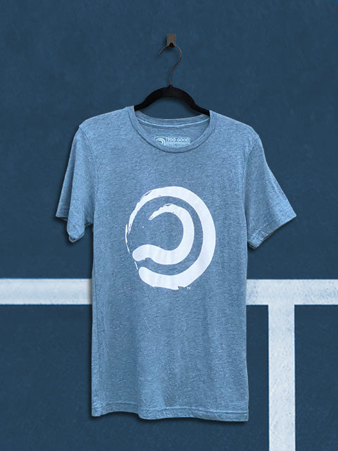 Denim blue colored t-shirt with a white ball mark graphic, hanging from a black hanger in front of a teal tennis court background.