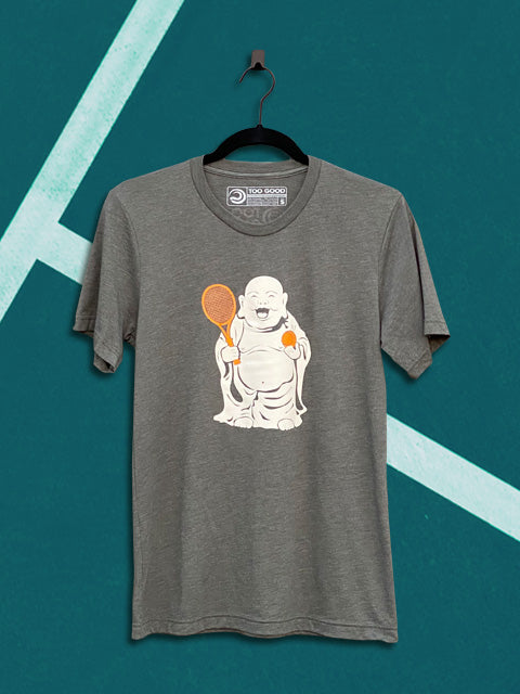 Military green graphic short-sleeve tennis t-shirt with a tan buddha graphic on the front holding an orange ball and racquet. The graphic tennis t-shirt is hanging on a black hanger in front of a teal tennis court background.