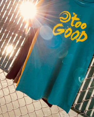 Teal short-sleeve t-shirt with bright yellow Too Good graphic hanging from a white hanger on a fence in front of a tennis court. A sun flare is bursting from behind the shirt, coming through the slats in the fence.