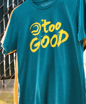 Close up of a teal short-sleeve t-shirt with bright yellow Too Good graphic hanging from a white hanger on a fence in front of a tennis court. Background is out of focus.