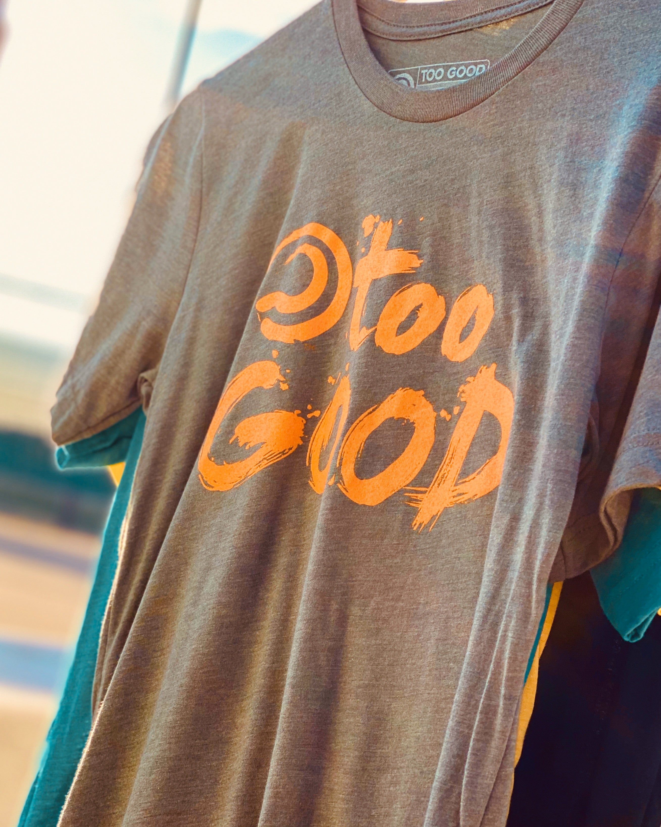 Close up a military green short-sleeve t-shirt with orange Too Good graphic hanging in front of a tennis court. The court is out of focus in the background.