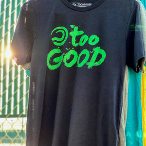 Close up of a black short-sleeve Too Good t-shirt with green graphic handing from a fence at a tennis court. Sun is shining through the fence.