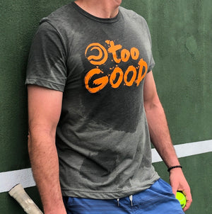 Shot of a tennis player from the neck down leaning against a practice wall, holding a ball and racquet and wearing a military green short sleeve t-shirt with orange Too Good graphic that has sweat marks.