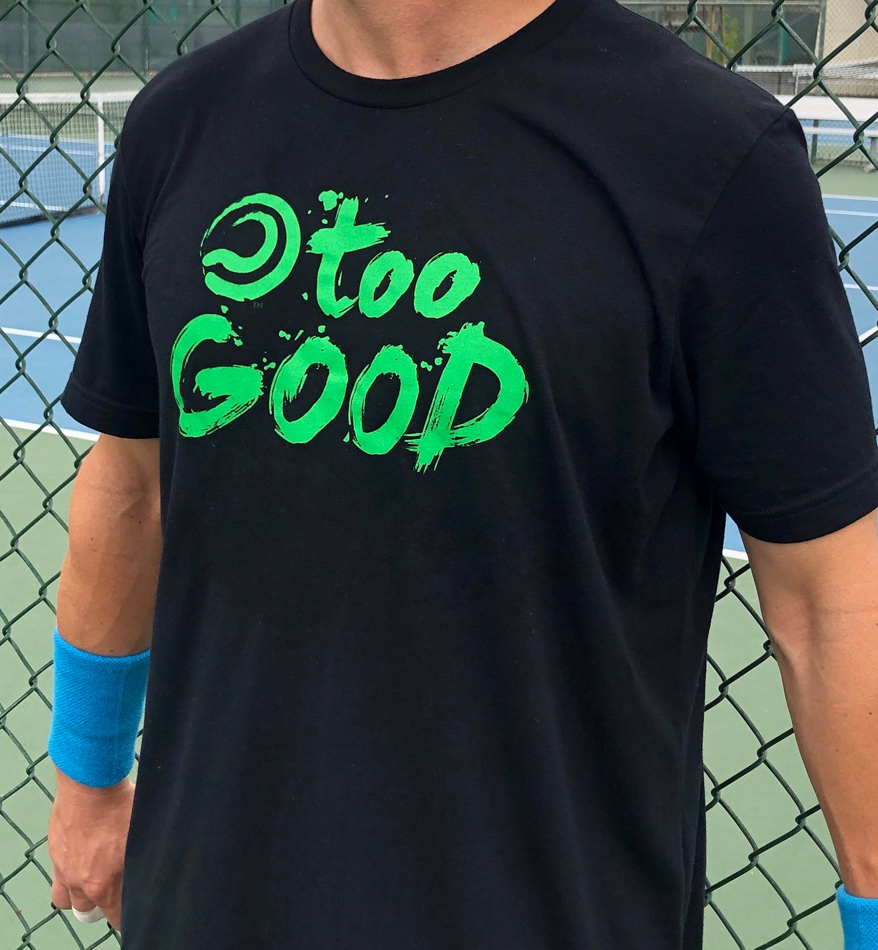 Shot of a tennis player's torso while wearing Black shortsleeve t-shirt with green Too Good graphic in front of a fence at a tennis court. Wearing blue wrist bands.