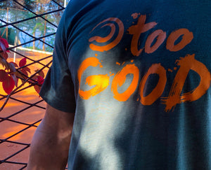 Close up of a tennis player in a military green short-sleeve t-shirt with orange Too Good graphic. He is in front of a fence with vines and leaves growing on it that surrounds a clay tennis court.