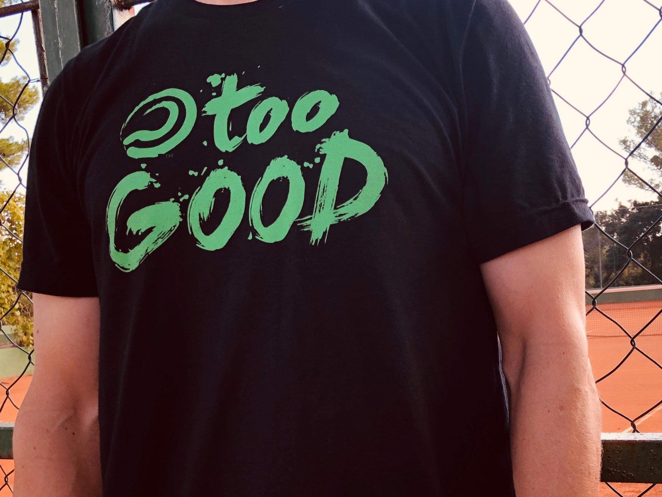 Shot of a tennis player's torso while wearing Black shortsleeve t-shirt with green Too Good graphic in front of a fence in front of a clay tennis court