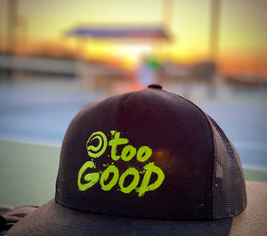 Too Good black trucker hat with mesh back and a snapback with a lime green Too Good graphic sitting on a bench in front of a blurred out blue tennis court background at sunrise with a beautiful orange and yellow glow.