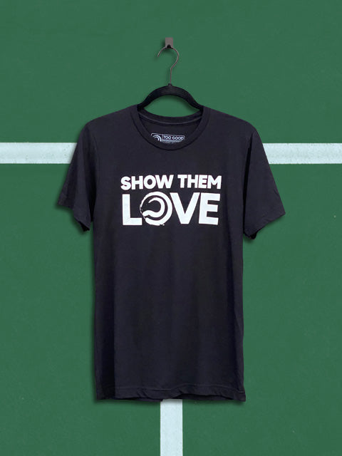 Black Too Good tennis t-shirt with an white graphic that says show them love, hanging from a black hanger in front of a green tennis court background.