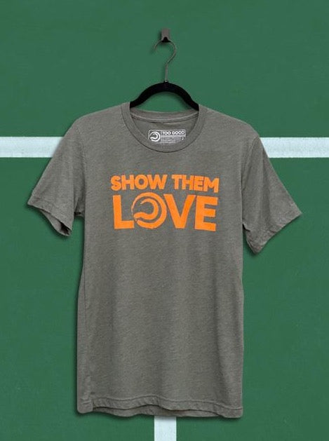 Denim military green colored t-shirt with an orange graphic that says show them love, hanging from a black hanger in front of a green tennis court background.