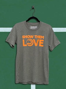 Denim military green colored t-shirt with an orange graphic that says show them love, hanging from a black hanger in front of a green tennis court background.