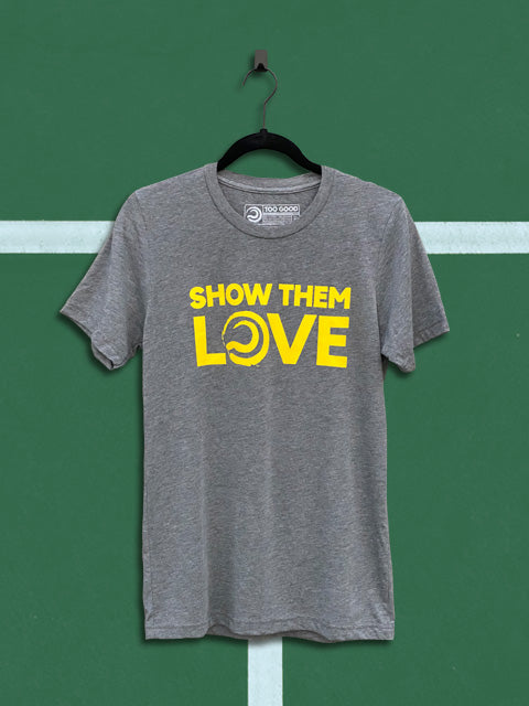 Heather grey shirt with yellow show them love graphic that has a too good ball where the o is in love hanging on a black hanger over a green court background