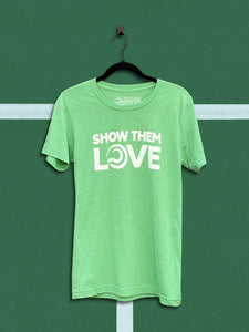 Green t-shirt with an white graphic that says show them love, hanging from a black hanger in front of a green tennis court background.