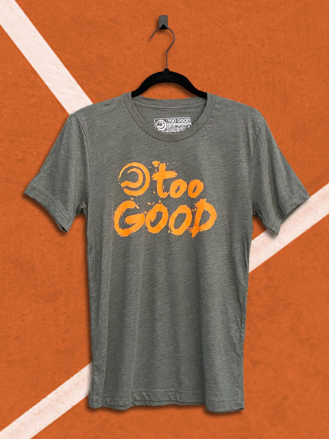 Military green t-shirt with an orange graphic that says too good with a ball mark, hanging from a black hanger in front of an orange tennis court background.