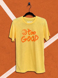 Yellow t-shirt with an orange graphic that says too good with a ball mark, hanging from a black hanger in front of an orange tennis court background.