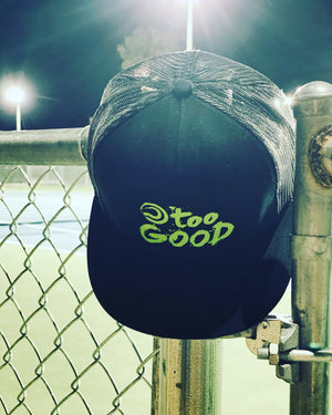 Too Good black trucker hat with mesh back and a snapback with a lime green Too Good graphic hanging on a fence at night under the lights with a blue and green tennis court in the background.